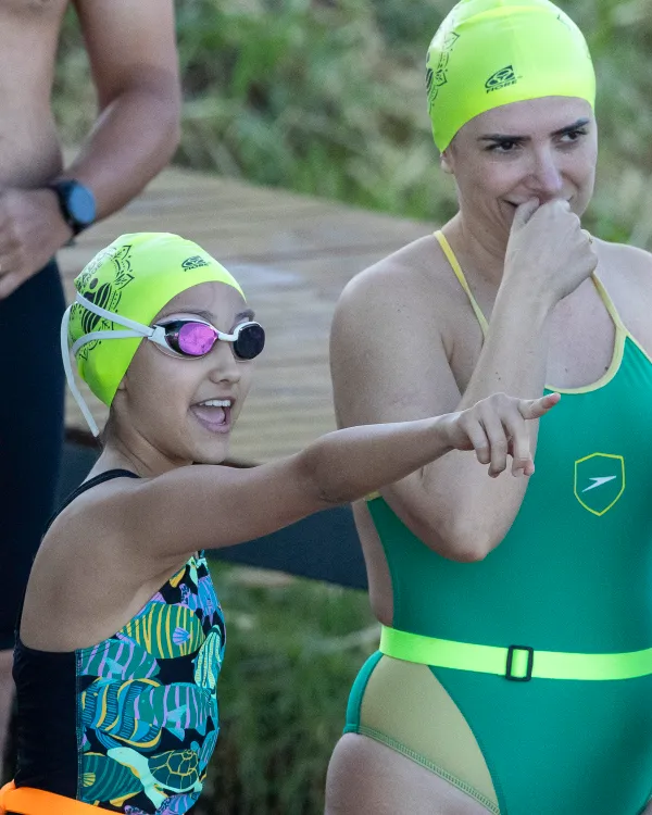 Menina de touca verde-limão conversa e ri com outra nadadora na beira do deque, antes de entrar na água