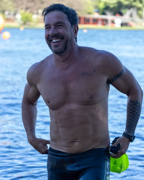 Nadador sai da água sorrindo, sem camisa, com a boia de segurança verde-limão na mão